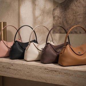 May include: Five structured handbags arranged in a row, featuring a single top handle and a central metal turn-lock closure. The bags are available in light pink, black, cream, dark brown, and tan, each with a textured finish and curved silhouette.