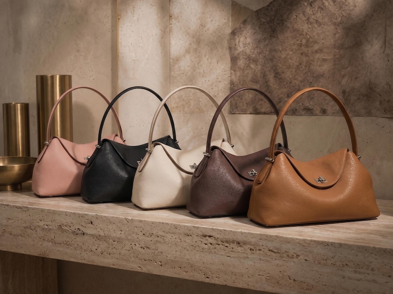 May include: Five structured handbags arranged in a row, featuring a single top handle and a central metal turn-lock closure. The bags are available in light pink, black, cream, dark brown, and tan, each with a textured finish and curved silhouette.