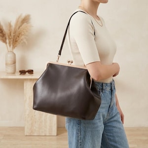 May include: A dark brown leather shoulder bag with a structured frame, a gold-toned metal kiss-lock closure, and a matching dark brown leather strap. The bag features a smooth, matte finish and a wide, rounded base for storage capacity.