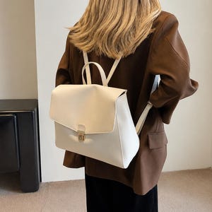 Puede incluir: Una mochila de color crema con solapa plegable y cierre de broche metálico en tono dorado. El bolso cuenta con un asa superior y correas ajustables, fabricado en un material liso y sólido con una forma rectangular estructurada.