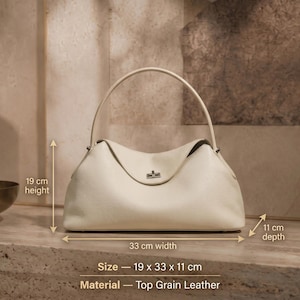 May include: A cream-colored top grain leather handbag with a single arched handle and a silver-tone turn-lock closure. The bag measures 19 cm in height, 33 cm in width, and 11 cm in depth. The text on the image specifies the size and material.