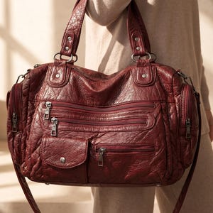 Puede incluir: Un bolso de mano color burdeos con textura arrugada, que cuenta con múltiples compartimentos exteriores con cremallera, un bolsillo pequeño con solapa y botón a presión, dos asas con remaches plateados y bolsillos laterales con cremalleras verticales.