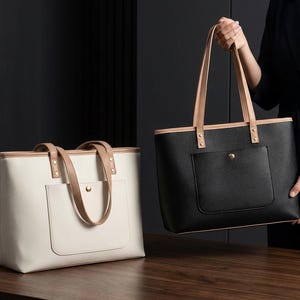 May include: Two rectangular tote bags, one in white and one in black, each featuring tan leather-like handles, tan trim along the top edge, and a single exterior front pocket secured with a gold-toned metal snap button closure.