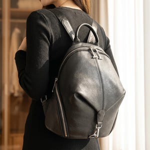 以下が含まれることがあります： 丸みを帯びたシルエットの黒いレザー製バックパック。トップハンドル、調節可能なショルダーストラップ、シルバーカラーの金属製ジッパーが特徴です。バッグの側面には縦型のジッパー付きポケットがあり、中央には縦の縫い目があしらわれています。
