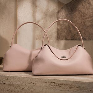 May include: Two light pink leather handbags with a soft, slouchy silhouette, a single rounded top handle, and a silver-toned metal turn-lock closure on the front. The bags feature a textured grain finish and a structured base with small feet.