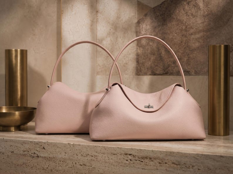 May include: Two light pink leather handbags with a soft, slouchy silhouette, a single rounded top handle, and a silver-toned metal turn-lock closure on the front. The bags feature a textured grain finish and a structured base with small feet.