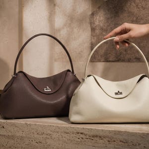 May include: Two structured handbags with a pebbled texture, one in dark brown and one in off-white. Each bag features a single arched handle, a curved top flap, and a silver-toned metal turn-lock closure centered on the front of the bag.