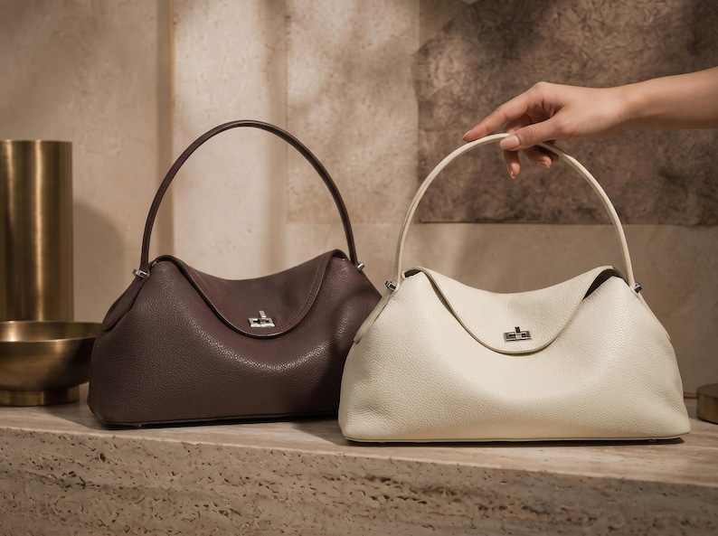 May include: Two structured handbags with a pebbled texture, one in dark brown and one in off-white. Each bag features a single arched handle, a curved top flap, and a silver-toned metal turn-lock closure centered on the front of the bag.