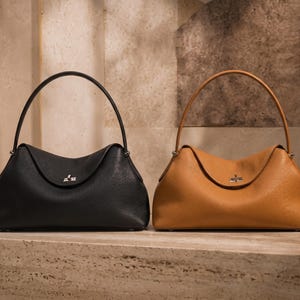 May include: Two structured handbags with a pebbled texture, one in black and one in tan. Each bag features a single arched handle, a curved top flap, and a silver-toned metal turn-lock closure centered on the front of the bag body.
