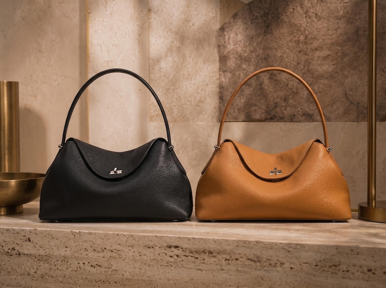 May include: Two structured handbags with a pebbled texture, one in black and one in tan. Each bag features a single arched handle, a curved top flap, and a silver-toned metal turn-lock closure centered on the front of the bag body.