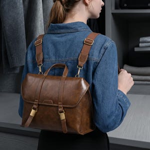 Puede incluir: Una mochila marrón de aspecto cuero con un asa superior y correas ajustables para los hombros. La bolsa cuenta con una solapa plegable asegurada por dos correas verticales con hebillas de metal dorado. El diseño incluye una base rectangular.