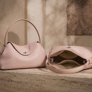 May include: Two pale pink leather handbags with a soft, pebbled texture. One bag is standing upright with a single arched handle and a silver-toned turn-lock closure, while the other is lying on its side, revealing a light-colored interior lining.