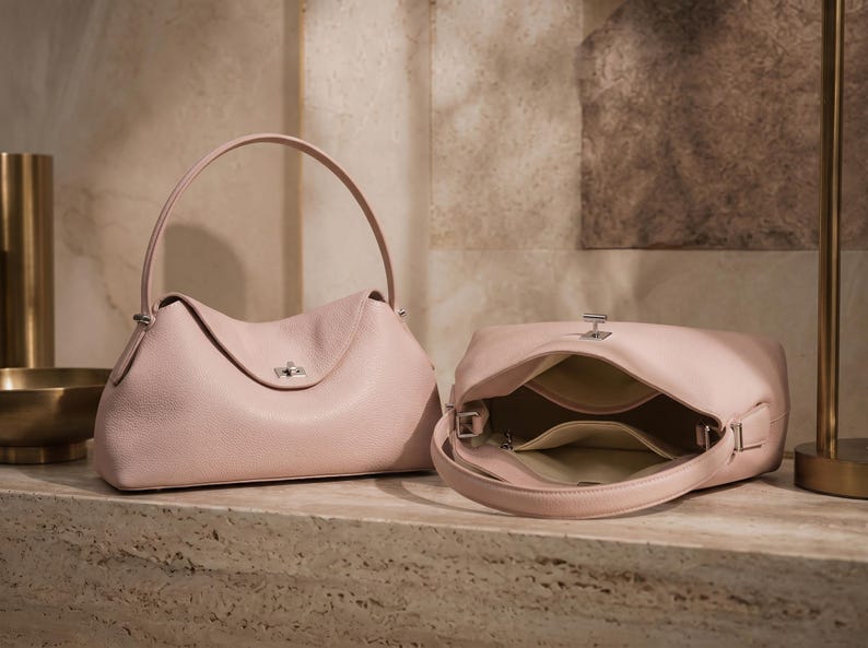 May include: Two pale pink leather handbags with a soft, pebbled texture. One bag is standing upright with a single arched handle and a silver-toned turn-lock closure, while the other is lying on its side, revealing a light-colored interior lining.