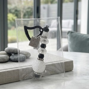 May include: A keychain featuring a black triangular clasp, a square silver charm engraved with the text BORN TO SHOP, and a series of alternating black, white, and clear textured beads with silver-toned accents, all enclosed in a clear plastic sleeve.