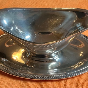 May include: A polished silver-tone gravy boat with a smooth, curved body and a wide pouring spout, resting on an integrated oval base plate that features a decorative rope-style texture along the outer edge of the rim.