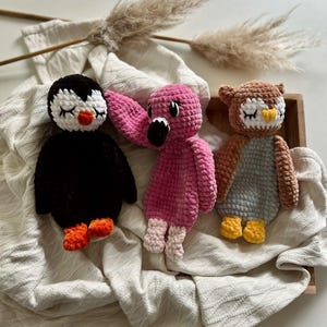 May include: Three handmade crochet plush toys featuring a black and white penguin with orange feet, a bright pink flamingo with a black beak, and a brown and gray owl with a yellow beak. Each stuffed animal has closed eyes and soft texture.