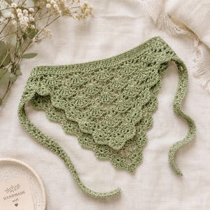 May include: A sage green triangular bandana featuring a delicate crochet shell stitch pattern with scalloped edges and long ties on each side. The accessory is crafted with a fine yarn texture and displays a consistent light green color throughout.