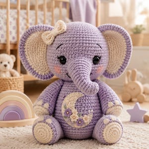 May include: A lavender crochet elephant stuffed animal featuring large ears with cream-colored centers, a small cream bow on the head, and a cream-colored crescent moon appliqué on the chest decorated with small purple and white flowers.