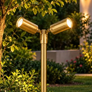 May include: A dual-head outdoor landscape light fixture with a metallic gold finish. The fixture features a vertical pole supporting two adjustable cylindrical spotlights that emit a warm, bright glow, positioned in a garden setting.