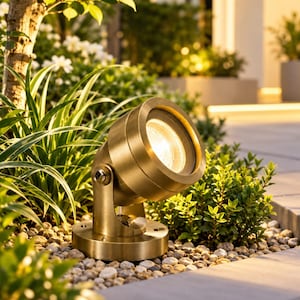 Puede incluir: Un foco de exterior cilíndrico de latón con cabezal giratorio ajustable y base circular. La luminaria presenta un acabado metálico cálido y está colocada entre plantas verdes de jardín y grava clara, emitiendo una luz brillante.