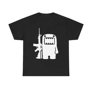 Może przedstawiać: Czarny t-shirt z krótkim rękawem z białym nadrukiem graficznym na klatce piersiowej. Grafika przedstawia stylizowaną białą postać z szerokimi, zębatymi ustami, stojącą obok szczegółowej ilustracji karabinu szturmowego.