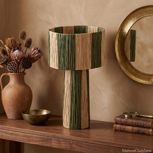 Può includere: Una lampada da tavolo con base cilindrica e un ampio paralume cilindrico abbinato, entrambi rivestiti da strisce verticali alternate di fibre color beige naturale e verde scuro, che conferiscono un aspetto strutturato e organico.
