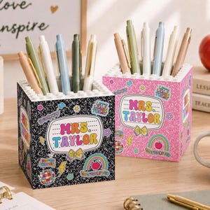 Puede incluir: Dos portalápices cuadrados con borde superior tipo bloque, uno negro y otro rosa, ambos con patrón moteado y pegatinas que dicen "MRS. TAYLOR" junto a iconos como arcoíris, lazos y bolas de discoteca, llenos de varios bolígrafos.