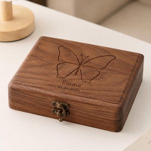 Puede incluir: Una caja rectangular de madera marrón oscuro con tapa abatible que presenta un diseño de mariposa grabado. La tapa está personalizada con el nombre Emma y la fecha 05.20.2024. La caja incluye un pequeño cierre de metal de estilo latón.