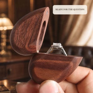 Può includere: Un portaanelli in legno marrone scuro con coperchio incernierato, contenente un anello in argento con una pietra chiara a taglio quadrato e un alone di pietre più piccole. L'etichetta rettangolare in alto a destra riporta il testo READY FOR THE QUESTION.