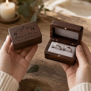 May include: A dark wood ring box with a hinged lid, featuring a laser-engraved mountain and pine tree design, the names James & Olivia, and the date 04.20.2026. The open box interior has a grey velvet insert holding two silver-toned rings.