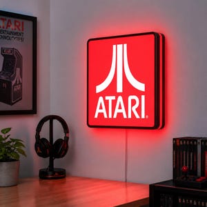 May include: A square wall-mounted LED sign featuring the white Atari logo and text on a vibrant red background. The sign emits a bright red glow from its edges, creating a luminous display that illuminates the surrounding wall space.