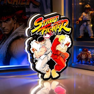 May include: A Street Fighter LED sign featuring the yellow and red logo at the top with illustrations of Ryu in a white gi and Ken in a red gi standing back-to-back. The sign has a black border and emits a bright white light from the edges.