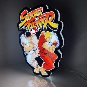 Può includere: Un'insegna da parete a LED retroilluminata con il logo Street Fighter in testo giallo e arancione sopra le illustrazioni di Ryu in gi bianco e Ken in gi rosso, entrambi intenti a eseguire attacchi energetici con effetti di luce blu brillante.