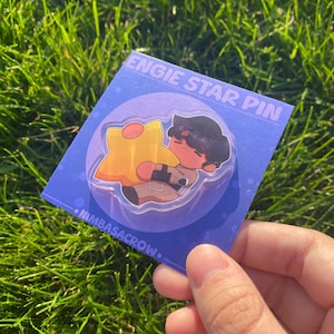 Peut inclure: Une broche en acrylique représentant un personnage de dessin animé aux cheveux foncés serrant une grande étoile jaune. La broche est fixée sur une carte de support carrée violette avec le texte ENGIE STAR PIN et NIMBASACROW imprimé en blanc.