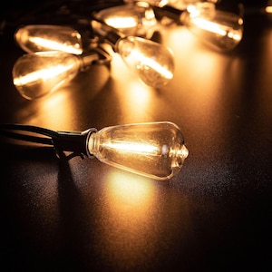 May include: A string of clear glass light bulbs with visible internal filaments emitting a warm, golden glow. The bulbs are connected by a black cord, with one bulb in the foreground clearly illuminated against a dark, reflective surface.