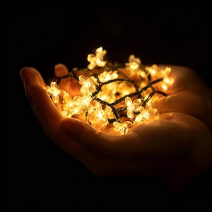 Puede incluir: Una cadena de luces LED con cubiertas translúcidas en forma de flor que emiten un brillo dorado y cálido. Las luces están conectadas por un cable fino y oscuro, creando un racimo de flores iluminadas que resplandecen sobre un fondo oscuro.