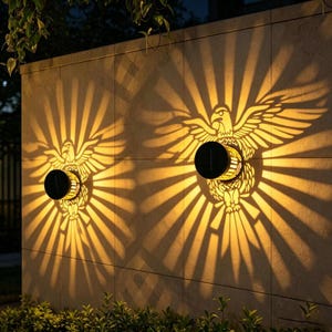 Könnte beinhalten: Zwei zylindrische Solar-Wandleuchten an einer Steinmauer, die jeweils ein helles, warmes Lichtmuster in Form eines Adlers mit ausgebreiteten Flügeln projizieren und einen strahlenden Sonneneffekt auf der flachen Oberfläche erzeugen.