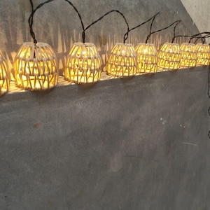 Op de afbeelding: Een lichtsnoer met kleine, klokvormige lampenkappen gemaakt van gevlochten, lichtgekleurde natuurlijke vezels. Elke kap bevat een warm gloeiende lamp, verbonden door een dunne donkere draad, wat een reeks verlichte lantaarns vormt.
