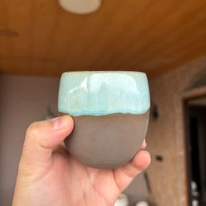 May include: A rounded ceramic cup featuring a smooth, matte dark brown base and a glossy, light turquoise glaze covering the upper portion. The vessel has a simple, handleless design with a curved silhouette and a clean, circular rim.