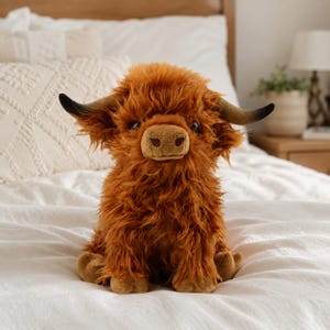 Op de afbeelding: Een pluche Highland-koe met lang, ruig, gemberbruin haar, een beige snuit en twee gebogen hoorns met donkere punten. Het knuffeldier zit rechtop en heeft donkere ogen en zachte, getextureerde vacht die het hele lichaam bedekt.