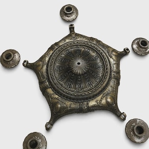 May include: An antique silver-toned metal centerpiece with a star-like shape, featuring intricate floral and geometric patterns. It has a central circular design and is accompanied by five smaller, matching candle holders.