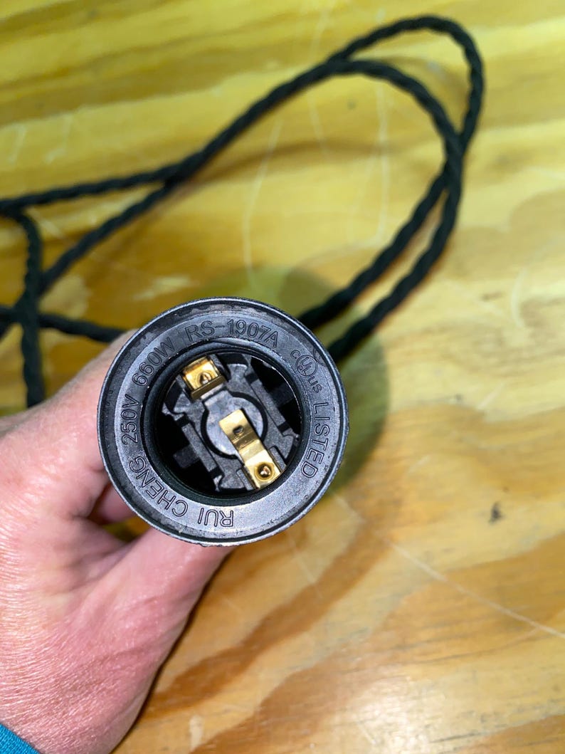 Op de afbeelding: Close-up van een zwarte lamphouder met goudkleurige metalen contacten. De lamphouder is gemarkeerd met "RS-1907A", "660W" en "250V". Een zwart, gevlochten elektrisch snoer is zichtbaar op de achtergrond. De lamphouder is gemarkeerd met "RUI CHENG" en "LISTED".