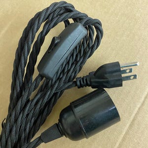 Könnte beinhalten: Schwarz geflochtenes Elektrokabel mit einem schwarzen Zwischenschalter, einem schwarzen Stecker und einer schwarzen Lampenfassung. Das Kabel ist eng gewebt. Komponenten sind für den elektrischen Gebrauch bestimmt. Kabellänge: ca. 100 cm.