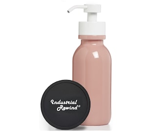 Puede incluir: Una botella dispensadora rosa con una bomba blanca y un posavasos redondo negro. El posavasos tiene las palabras "Industrial Rewind" en escritura blanca. La botella es de color rosa claro.