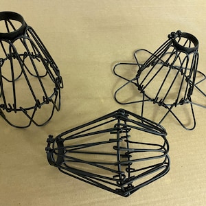 May include: Three black metal wire lamp shades in different shapes. One is cone-shaped, another is a starburst, and the third is a rounded cage. The shades are made of thin, black metal rods joined together.