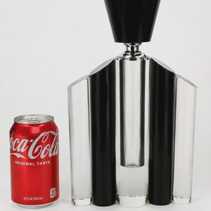 May include: A red Coca-Cola soda can with 12 fl oz (355 ml) text next to a tall, multi-faceted perfume bottle. The bottle features alternating clear and black vertical columns with a silver neck and a large, flared black cap on top.