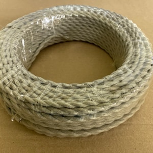 May include: A coiled beige electrical cable, wrapped in clear plastic. The cable has a braided texture and appears unused. The image is taken from a slightly elevated angle, showing the circular shape of the cable.
