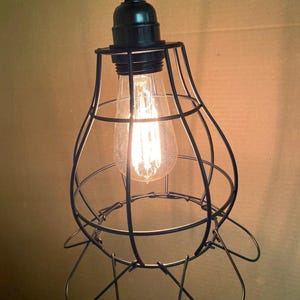 May include: A black metal pendant light fixture with a cage-like shade. The light bulb is visible inside the cage, emitting a warm, yellow glow. The fixture is suspended from a black cord.