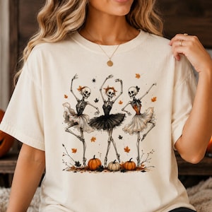Puede incluir: Una camiseta de manga corta de color crema con un estampado gráfico de tres esqueletos vestidos de bailarinas con tutús blancos, negros y naranjas. Los esqueletos bailan entre hojas de otoño dispersas y tres pequeñas calabazas en el suelo.