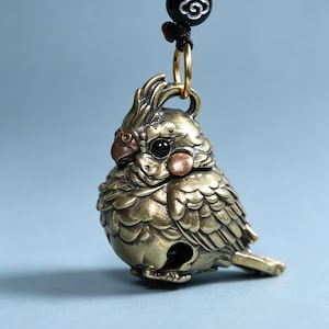 Peut inclure: Un pendentif en laiton en forme de calopsitte avec des textures de plumes détaillées, une petite crête et des yeux sombres. L'oiseau présente des accents cuivrés sur le bec et les joues, suspendu à un anneau doré et une perle noire à motif blanc.